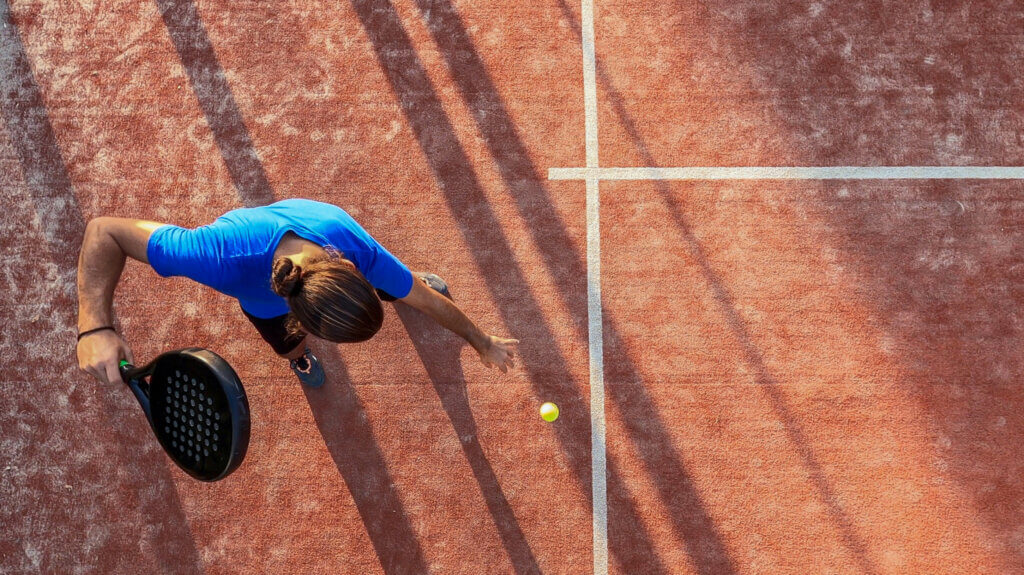 Top view of a paddle tennis player bouncing the ball for a serve on an outdoor court.